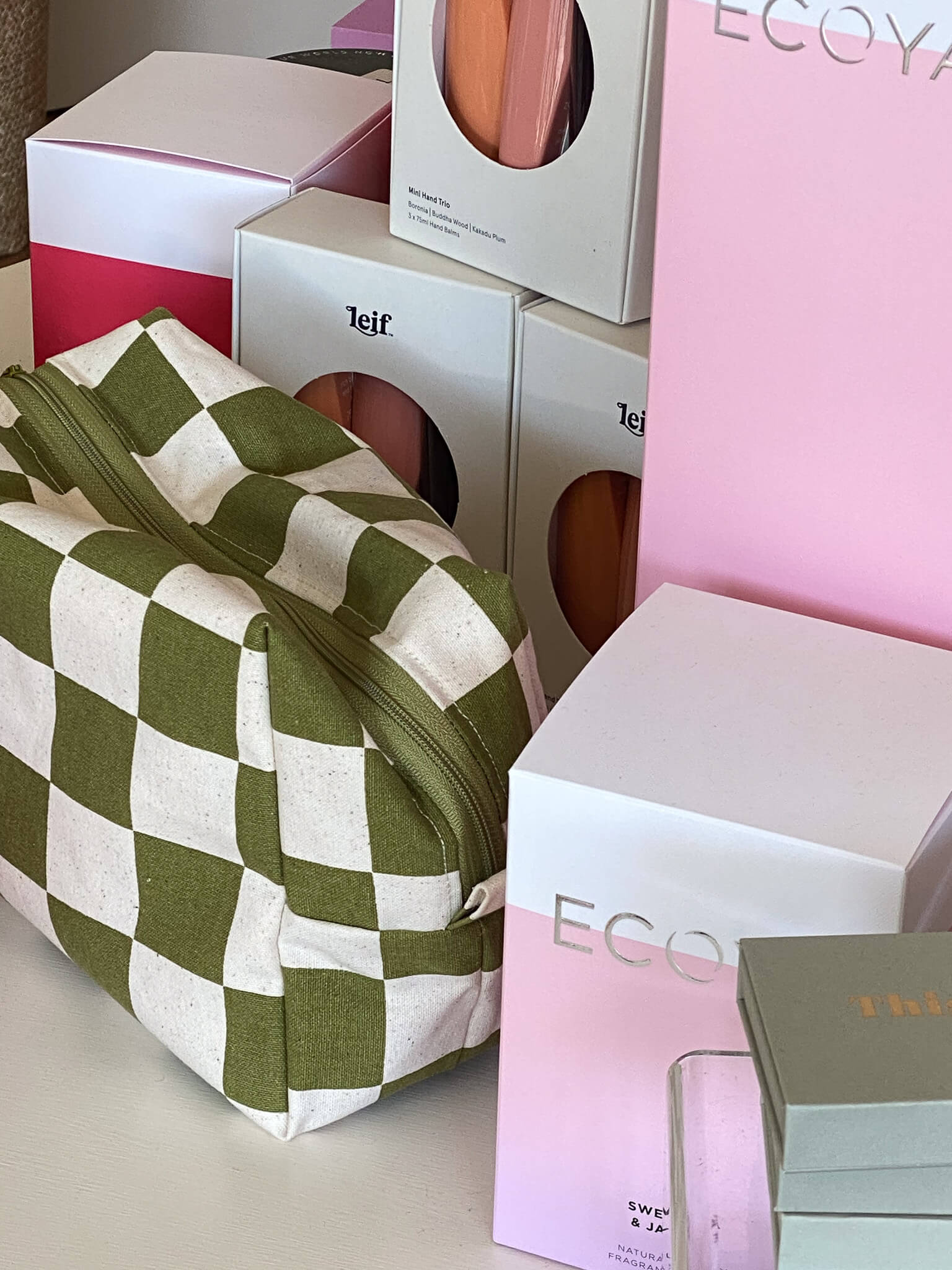 Green checkered pouch displayed alongside Ecoya and Leif product boxes on a white surface, featuring pastel pink and orange tones.
