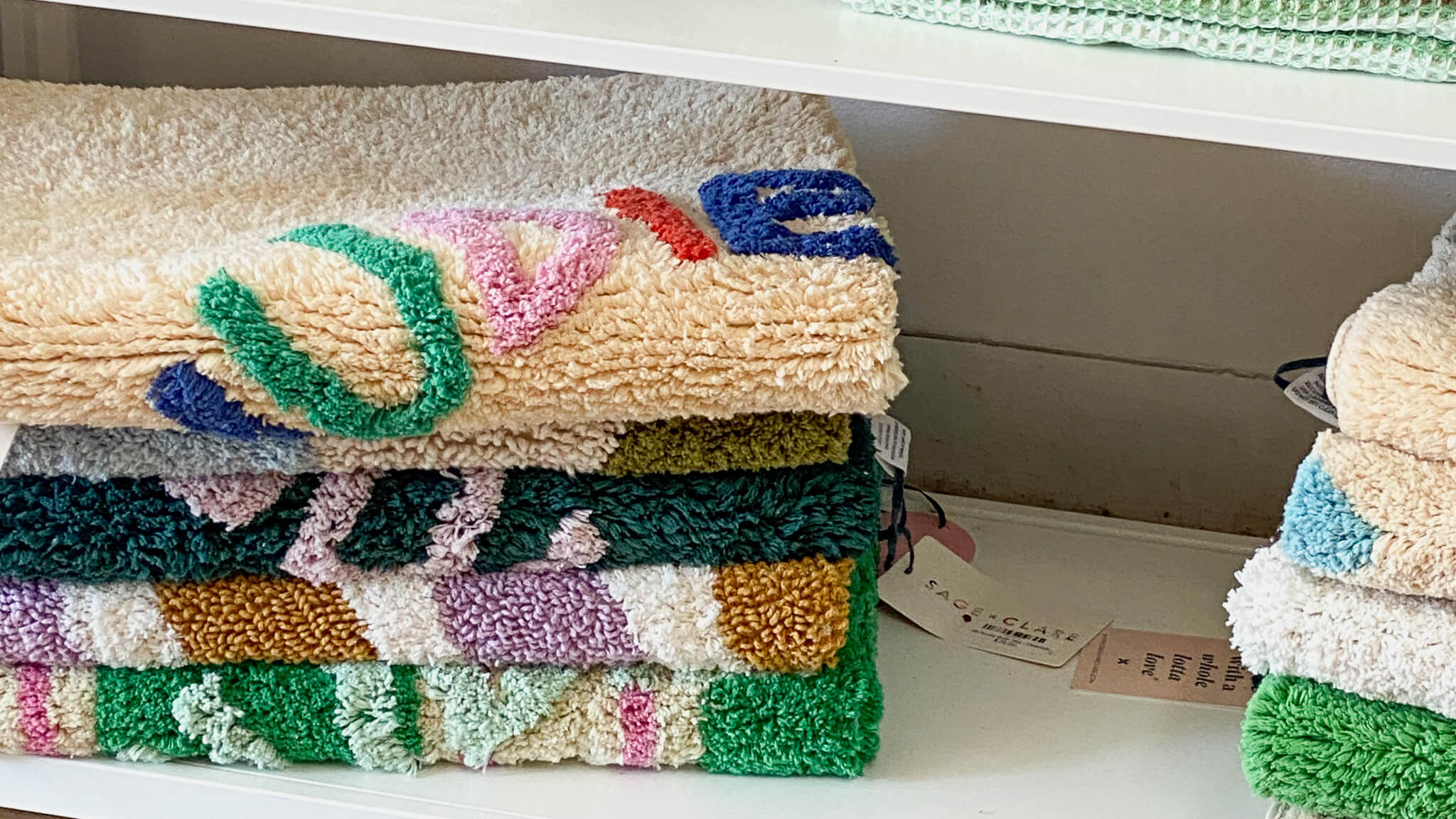 Colorful, patterned Sage + Clare towels neatly stacked on a white shelf, featuring soft textures and vibrant designs.